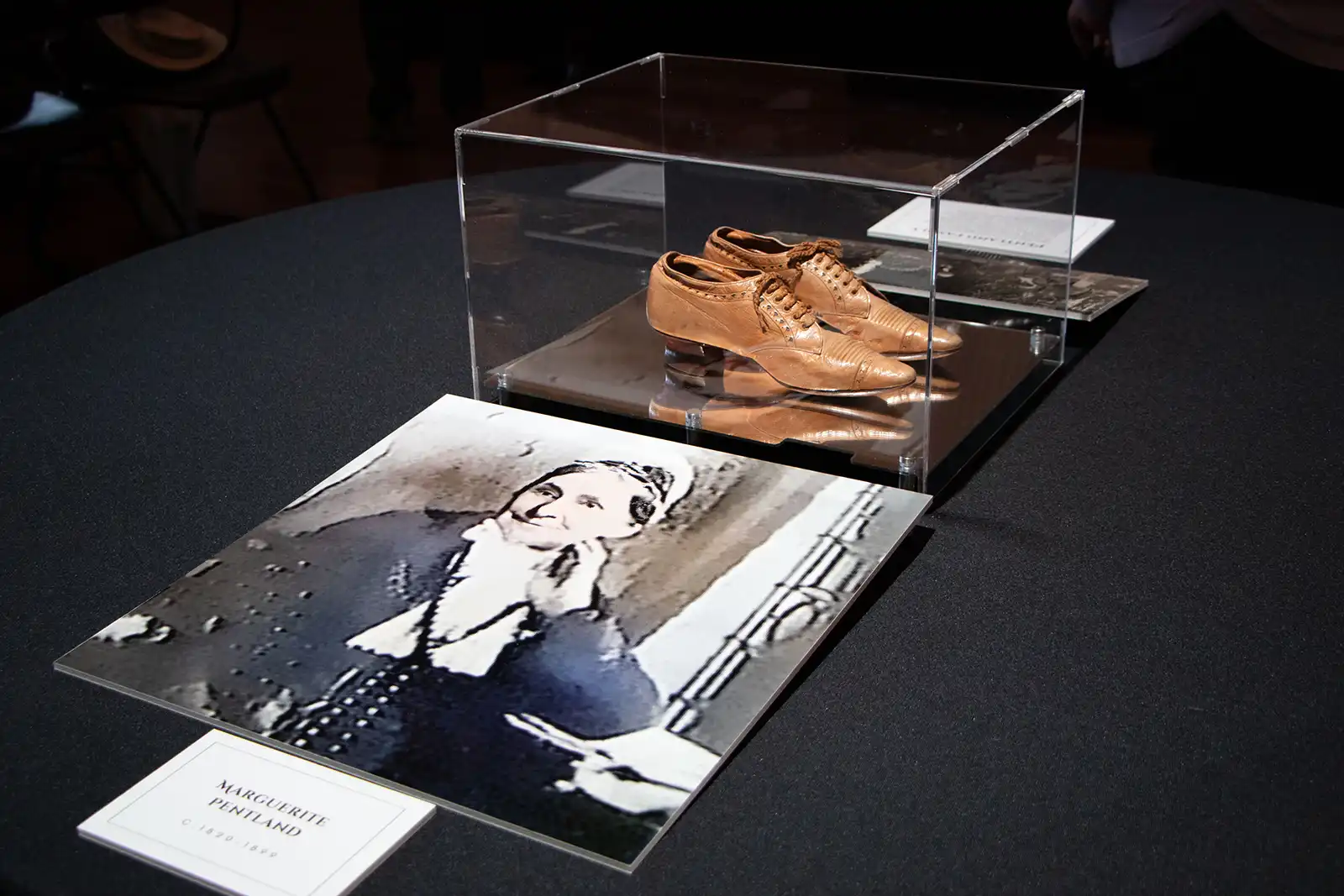 Photograph of shoes worn by Marguerite Pentland. Pictured in a glass case on a black tablecloth, with enlarged photo of Marguerite in front.